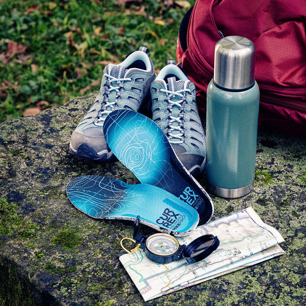 CURREX HIKEPRO insoles next to hiking accessories: shoes, thermos, backpack, compass and map sitting on moss-covered rock #1-wahle-dein-profil_low